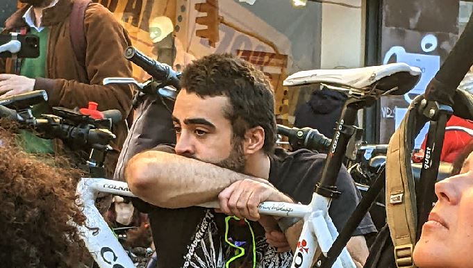 Ragazzo poggiato sulla sua bici, in mezzo a una folla, assorto